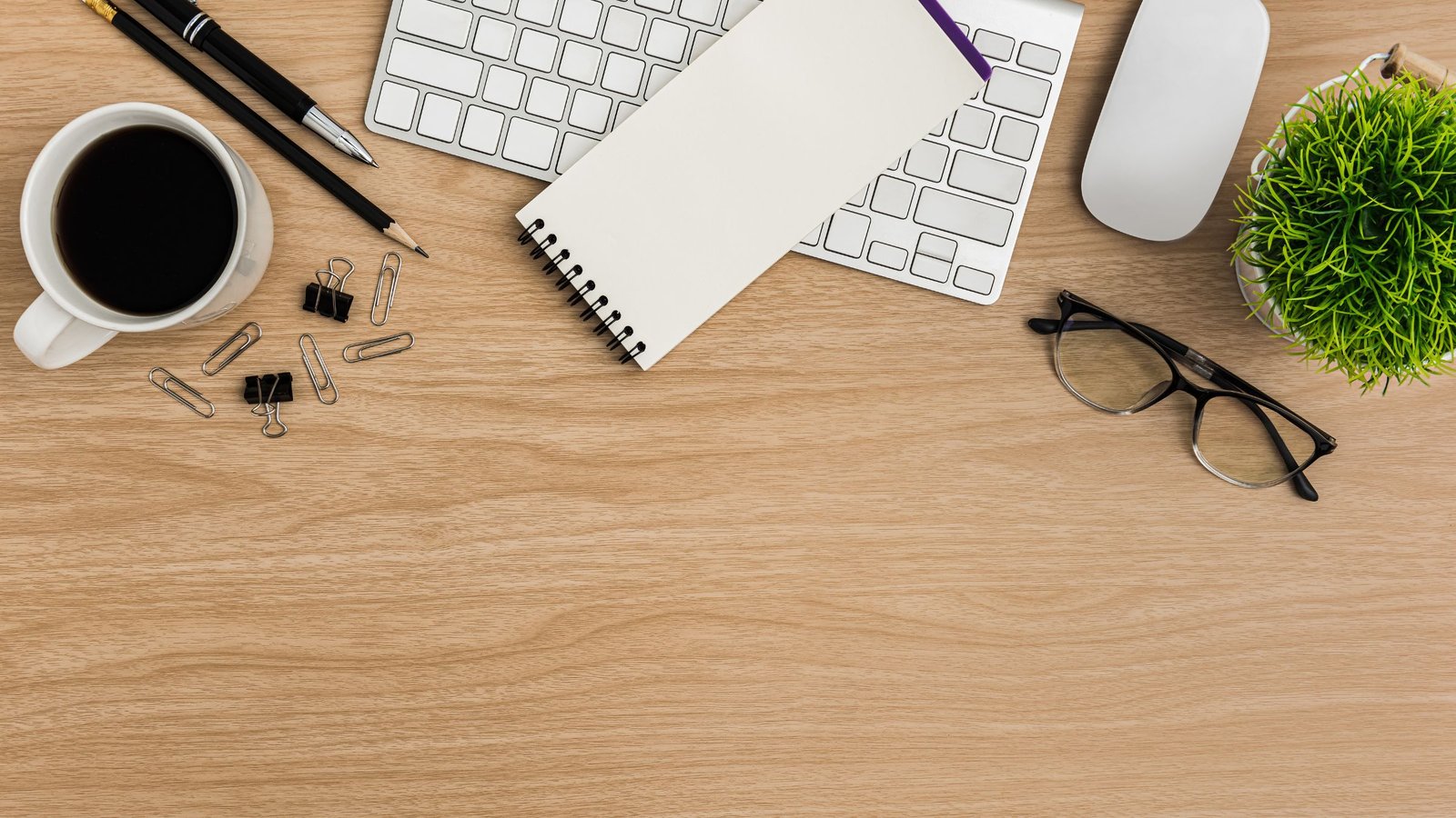 Wooden desk with a notebook, coffee cup, keyboard, glasses and paper clips